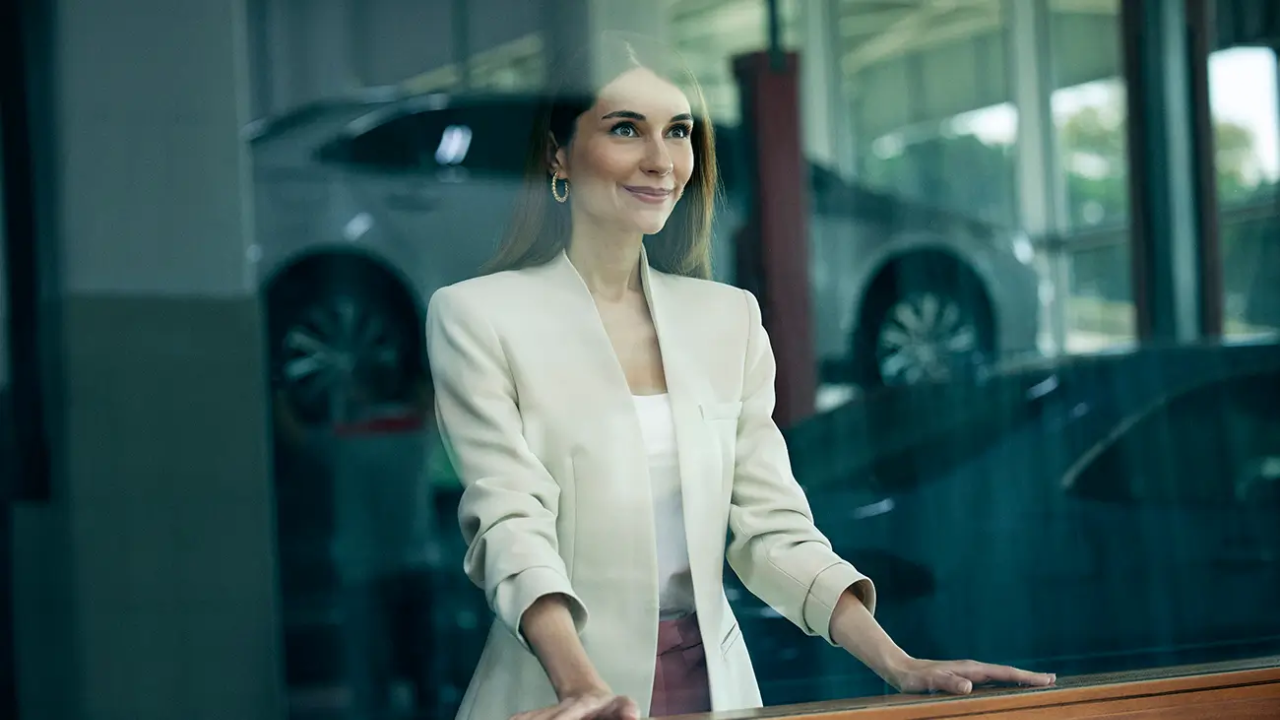 Arabaların olduğu bir alanda beyaz ceketli gülümseyen bir kadın