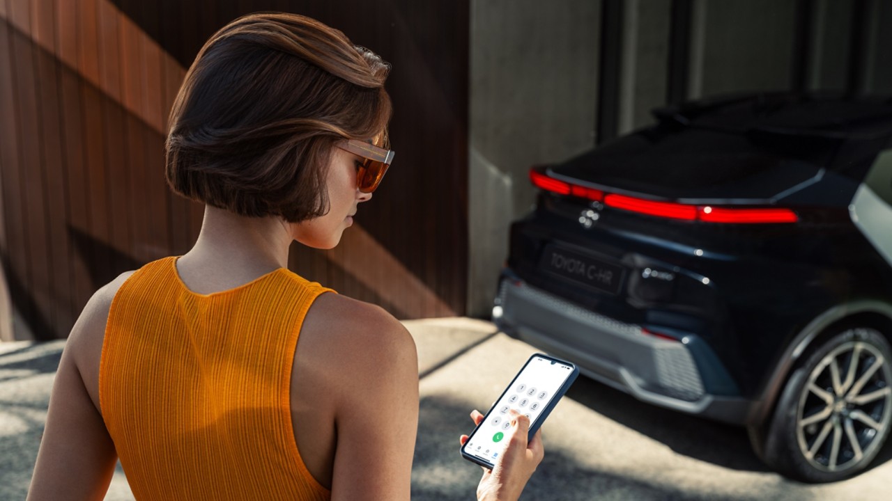 Toyota C-HR arkasında duran ve elindeki telefon ekranına bakan gözlüklü, kısa saçlı ve turuncu elbiseli bir kadın