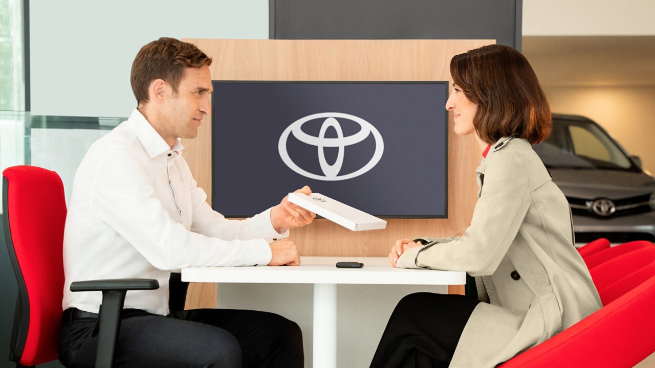 Toyota logosu önünde masada karşılıklı oturan bir kadın ve ona bir kitapçık uzatan bir adam